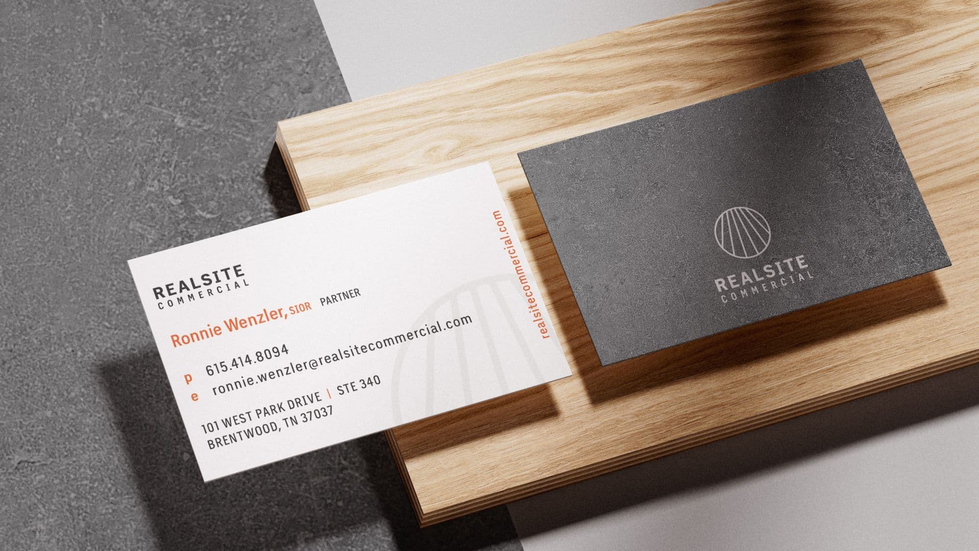 A high-angle photo shows two RealSite Commercial business cards arranged on a wooden block. The white card on the left displays contact information, while the dark gray card on the right features a white, circular logo and the company name.