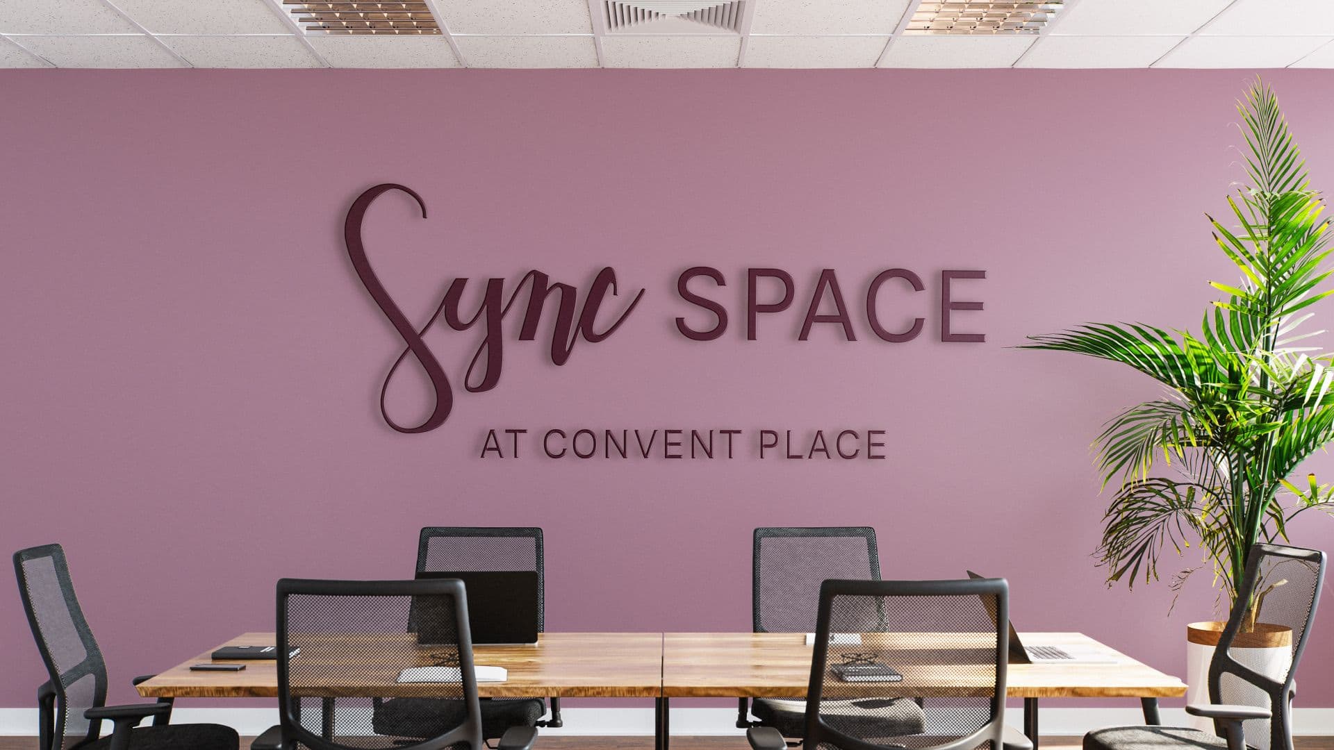 An interior shot of a meeting room shows a mauve-colored wall with the name Sync SPACE in three-dimensional, dark purple letters. Below this, the words AT CONVENT PLACE are visible in smaller lettering. A long wooden table with several black mesh chairs sits in the foreground, with a large potted plant to the right.